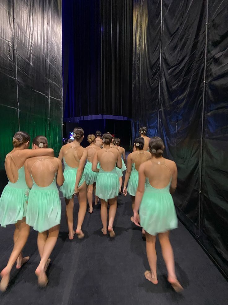 a group of young women in green dresses walking down a dark carpeted stage with their backs turned to the camera