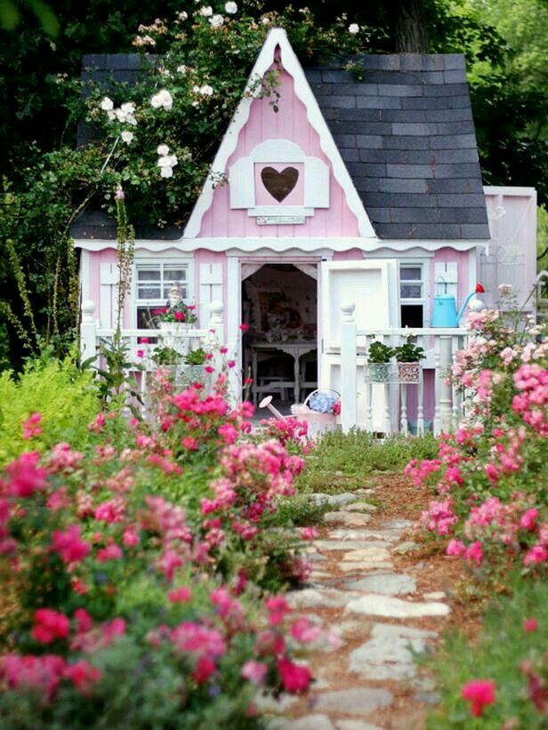 a small pink and white house in the middle of some flowers with a heart on it's window