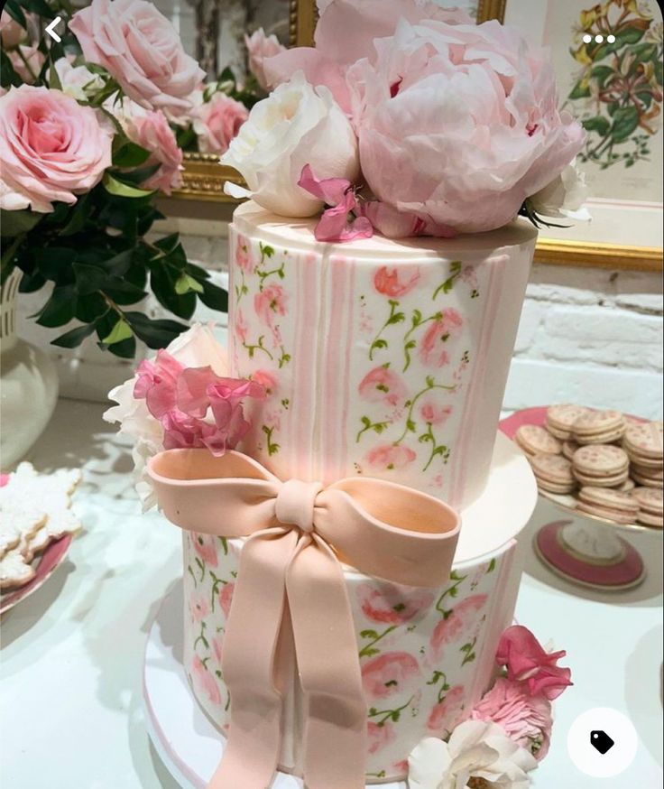 a three tiered cake decorated with pink flowers and ribbons on a table next to cookies