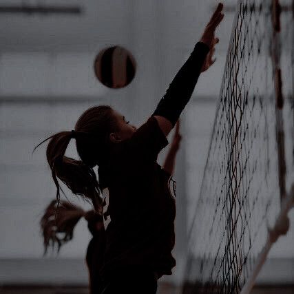 two girls are playing volleyball in an indoor court, one is reaching up to hit the ball with her racket