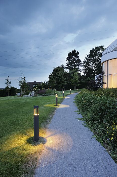 two lights that are on the side of a path near some bushes and grass in front of a building