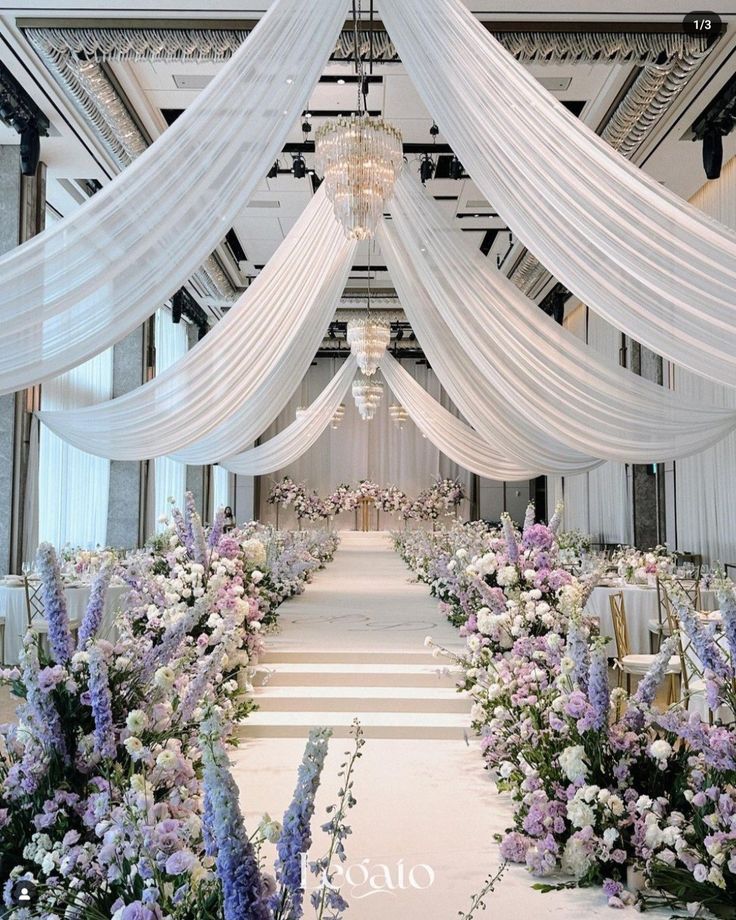 an aisle decorated with white drapes and purple flowers on either side is a chandelier that hangs from the ceiling