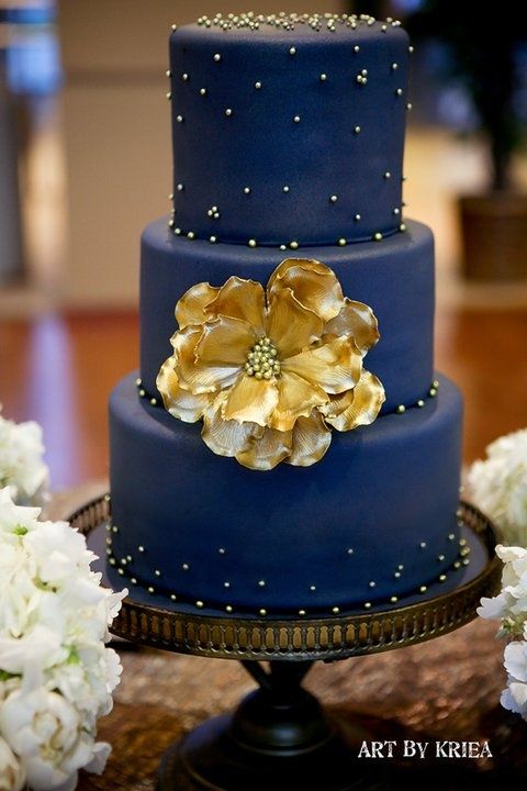 a three tiered blue cake with gold flowers on the top and bottom, sitting on a table