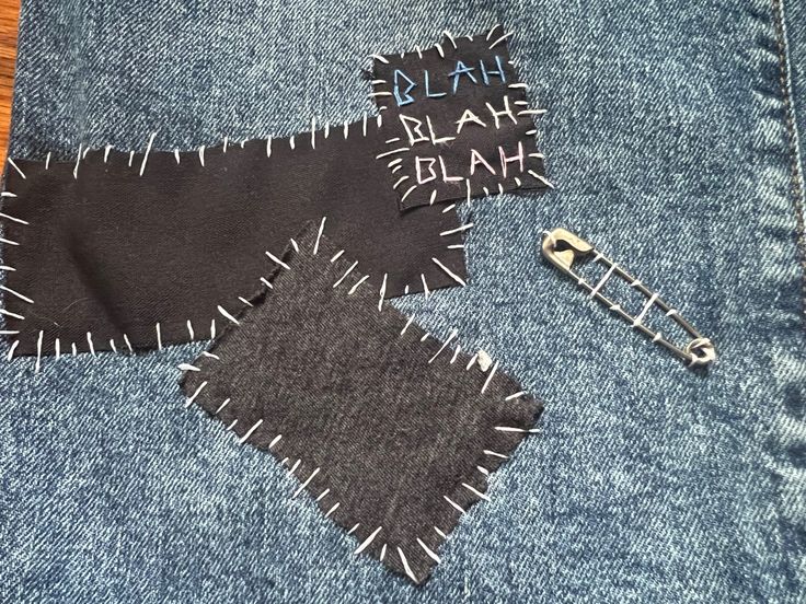 Black and grey fabric sewn onto blue jeans. A patch that reads blah blah blah written in blue, white, and pink is also sewn on. A safety pin is attached next to the 3 pieces of fabric. Punk Safety Pins Fashion, Pins On Pants, Sewn On Patches, Diy Punk Jeans, Patch Jeans Punk, Safety Pin Outfits, Punk Pins Diy, Punk Crafts Diy, Punk Pants Diy