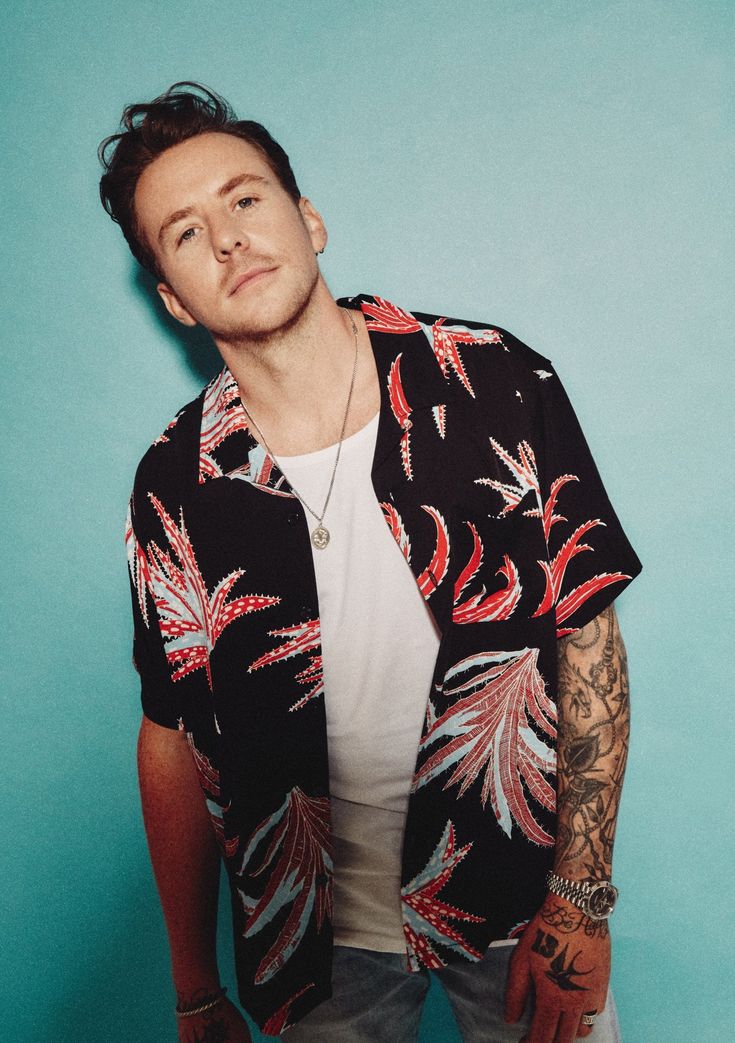 a man with tattoos standing in front of a blue wall wearing a black and red shirt