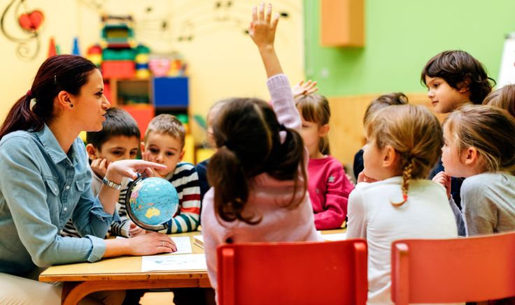 a woman sitting at a table in front of children with their hands up to the sky