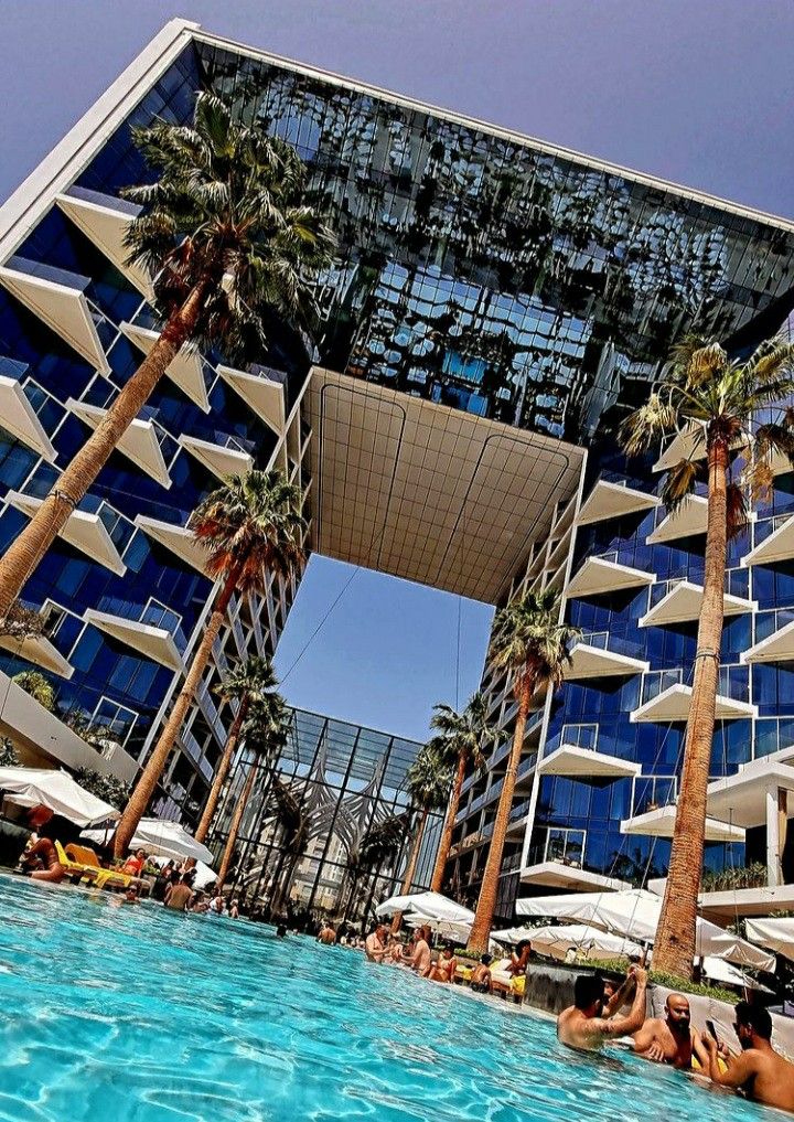 people are lounging in the swimming pool at an outdoor hotel with palm trees and blue glass windows