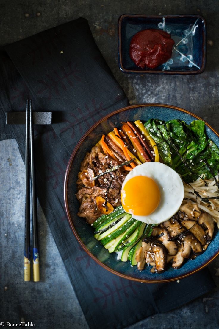 a plate with meat, vegetables and an egg on it next to chopsticks