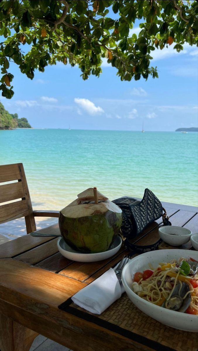 a plate of food is sitting on a table by the ocean with a coconut drink