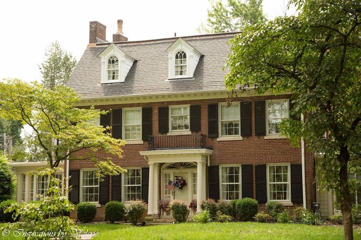 a large brick house with black shutters and white trim on the front door is surrounded by lush green trees