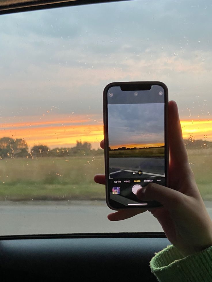 a person holding up a cell phone in front of a window with the sun setting