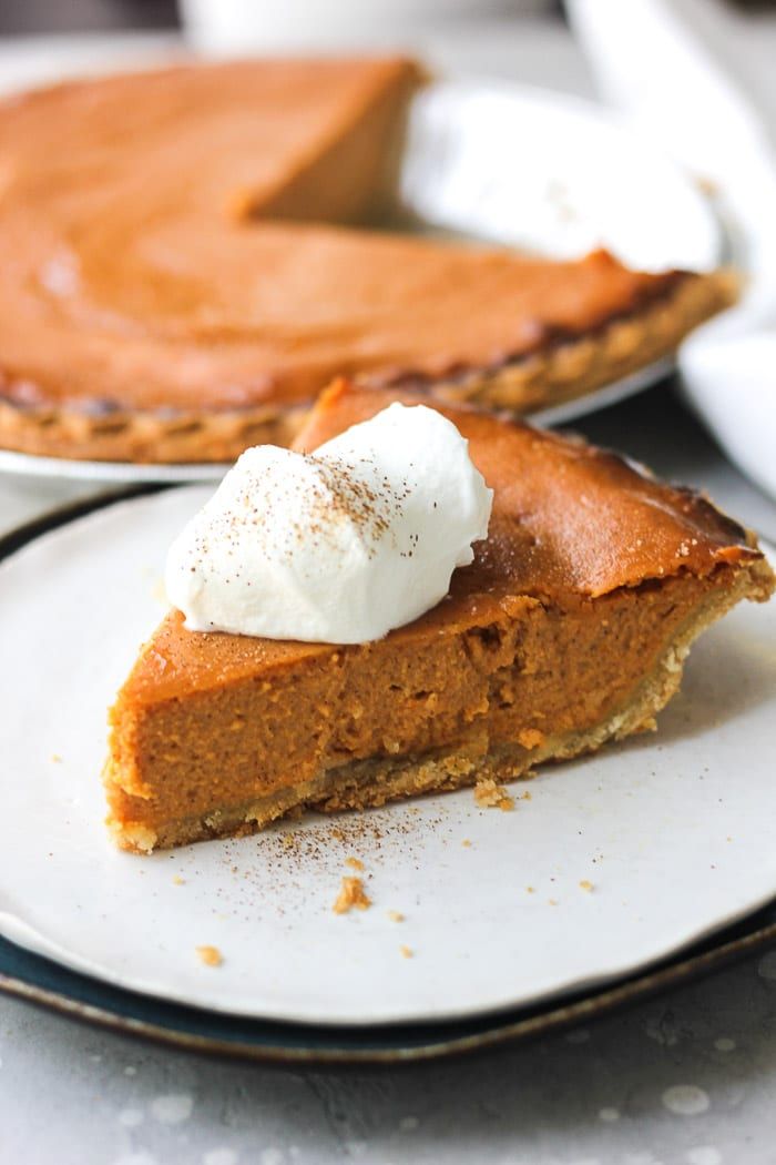 a slice of pumpkin pie with whipped cream on top sits on a plate in front of the rest of the pie