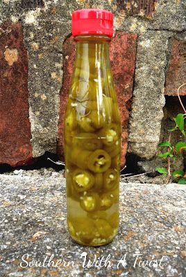 a glass bottle filled with green olives sitting on top of a cement ground next to a brick wall