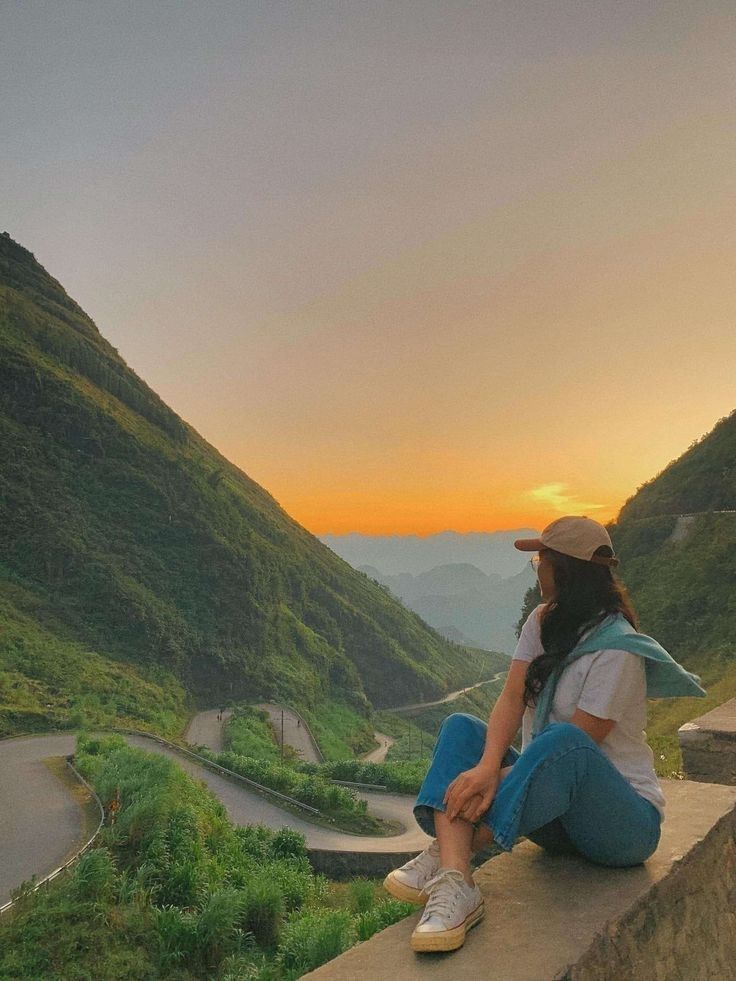 a woman sitting on top of a stone wall next to a lush green mountain range