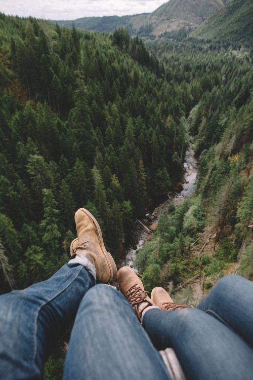 two people are sitting on the edge of a high cliff overlooking a river and forest