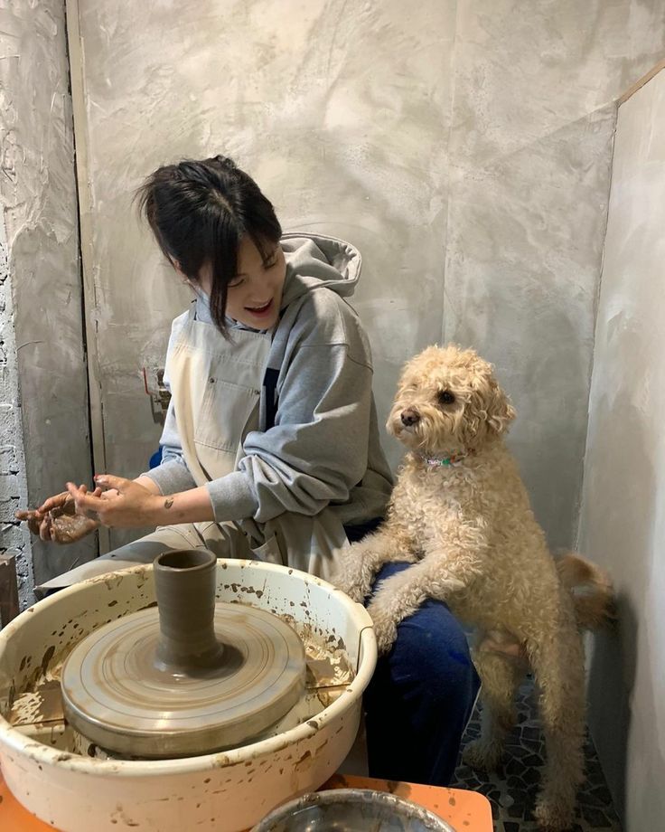 a woman sitting on the floor next to a pottery wheel with a dog in front of her