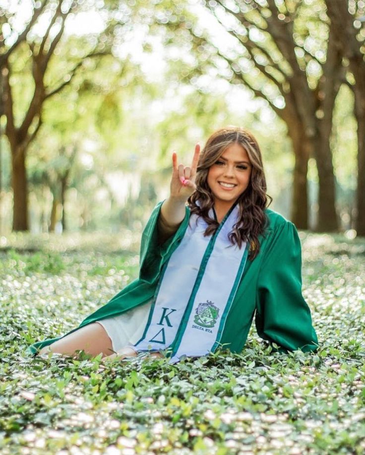 a woman sitting on the ground with her hand up in the air while wearing a green graduation robe