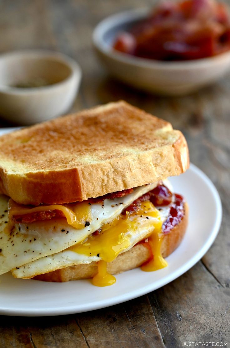 a breakfast sandwich with eggs, bacon and cheese on a white plate sitting on a wooden table