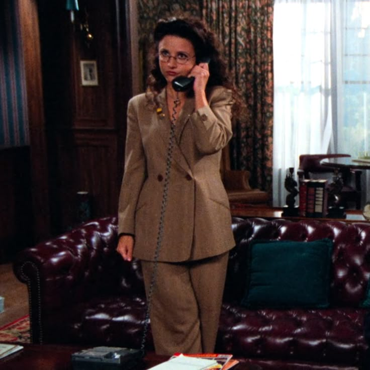 a woman in a suit talking on a cell phone while standing next to a couch