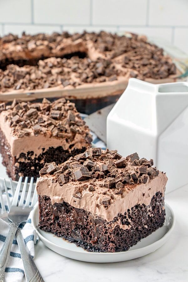 a slice of chocolate cake on a plate next to a fork and knife with the rest of the cake in front of it