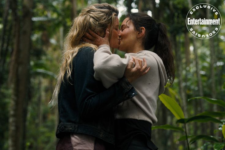 two women are kissing in the middle of a forest with trees and plants behind them