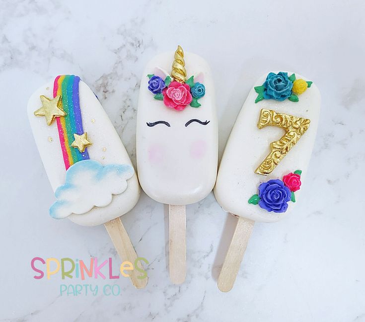 three ice cream pops decorated with unicorn faces and flowers on top of a marble surface