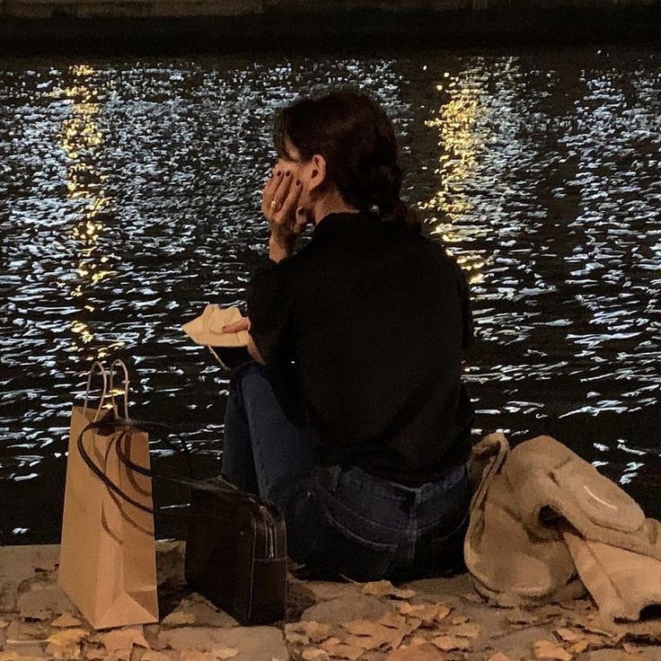 a woman sitting by the water with her hands to her face and shopping bags in front of her