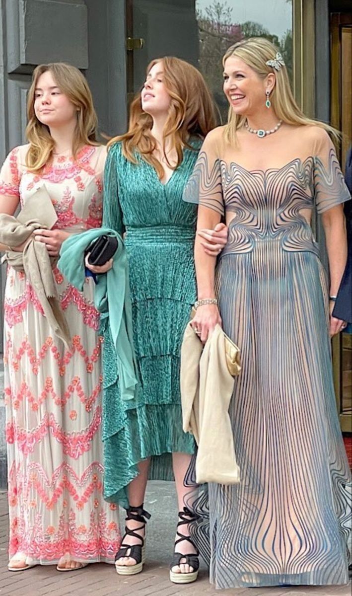 three women standing next to each other in front of a building wearing dresses and holding purses