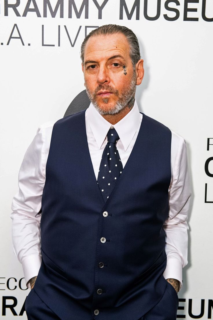 a man in a blue vest and tie standing next to a white wall with words on it