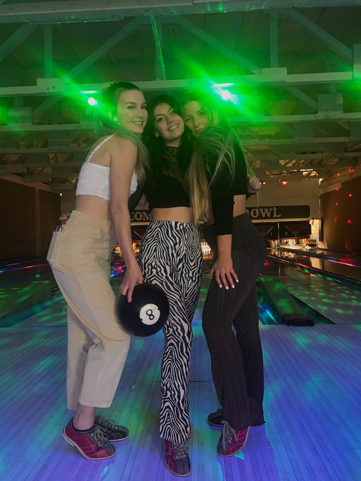 three women posing for the camera in front of bowling alley lanes with lights on them