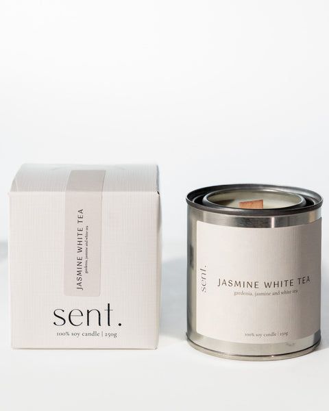 a candle in a tin next to a box on a white surface with the lid open