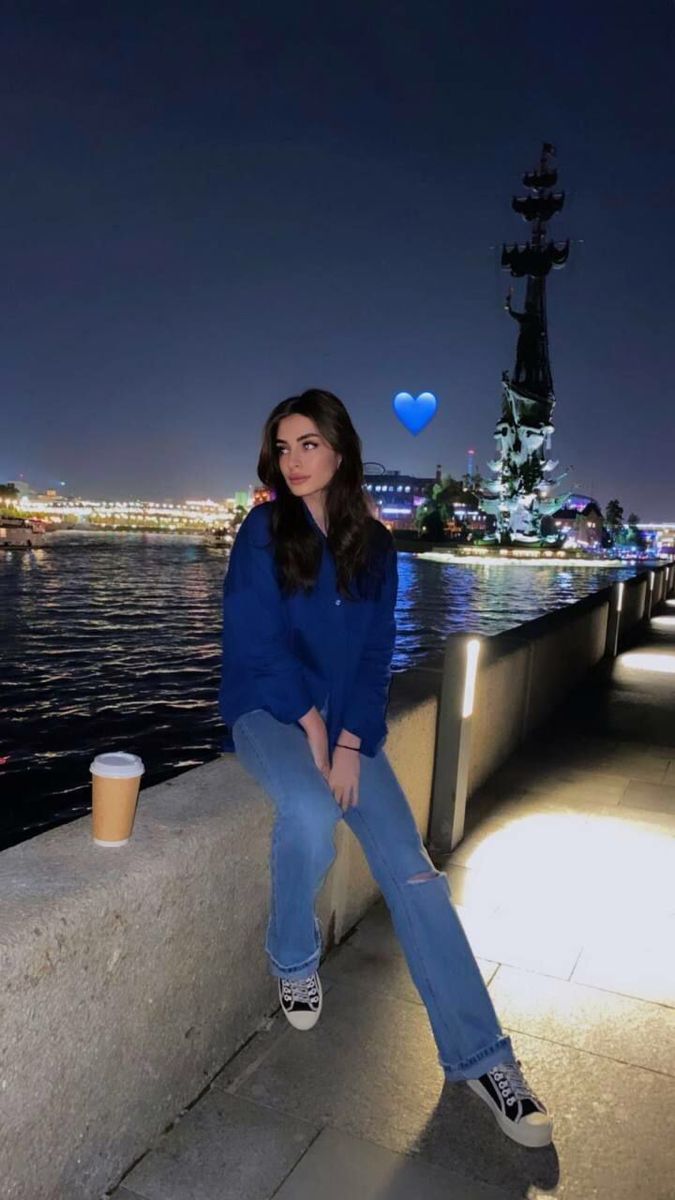 a woman sitting on a wall next to the water at night with balloons floating in the air