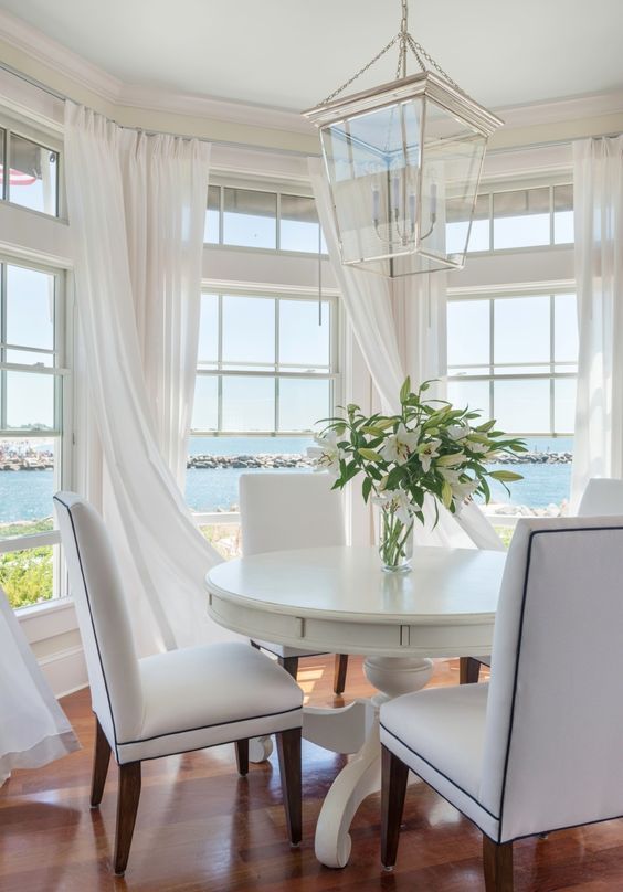 a dining room table with white chairs and a vase on top of it in front of two large windows