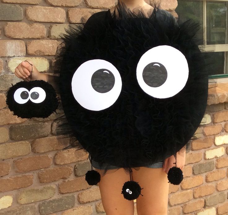 a woman standing next to a brick wall wearing a black furry ball with googly eyes on it