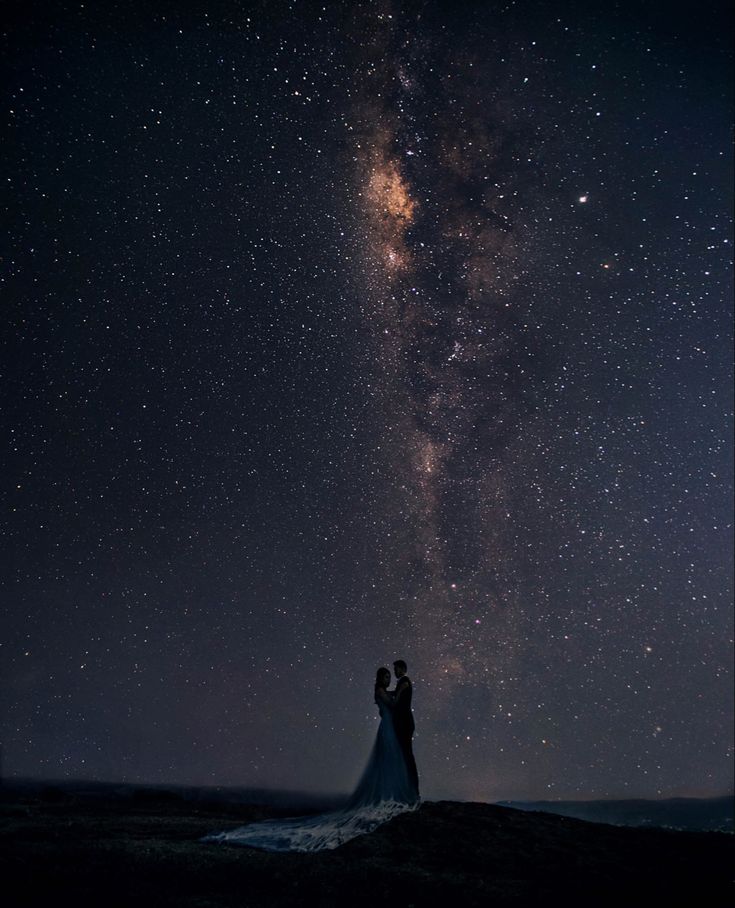 a bride and groom standing under the stars in the night sky with their arms around each other