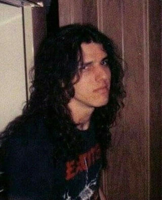 a man with long hair standing next to a refrigerator