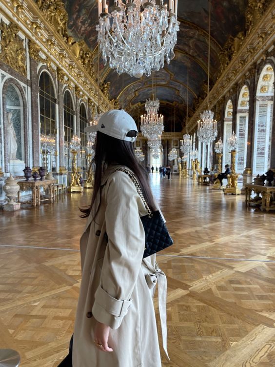 a woman wearing a white hat and trench coat in a large room with chandeliers