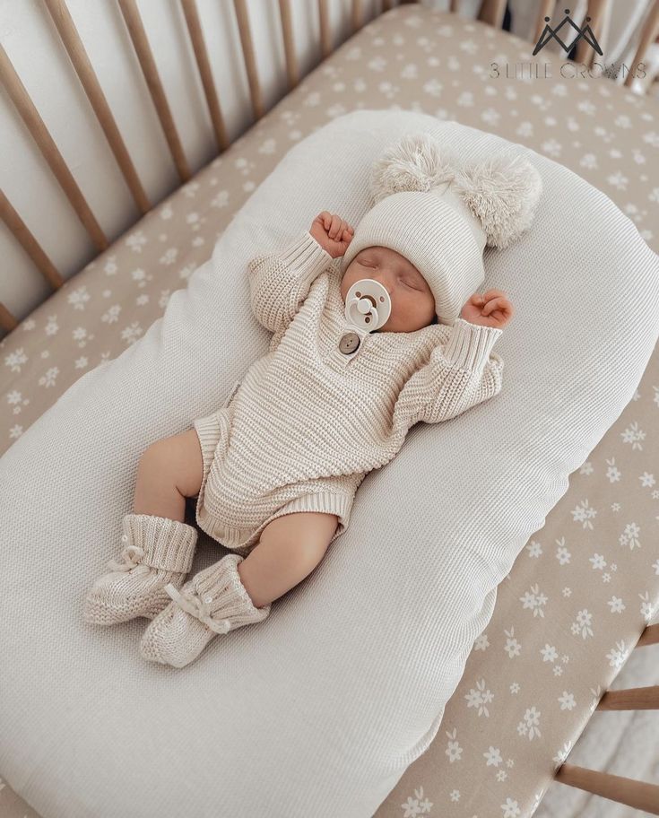 a baby laying in a crib wearing a white hat and sweater with a teddy bear on it