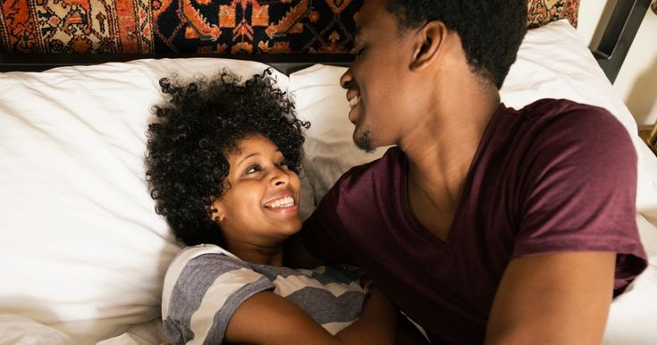 a man and woman laying in bed with pillows on top of each other, looking at each other