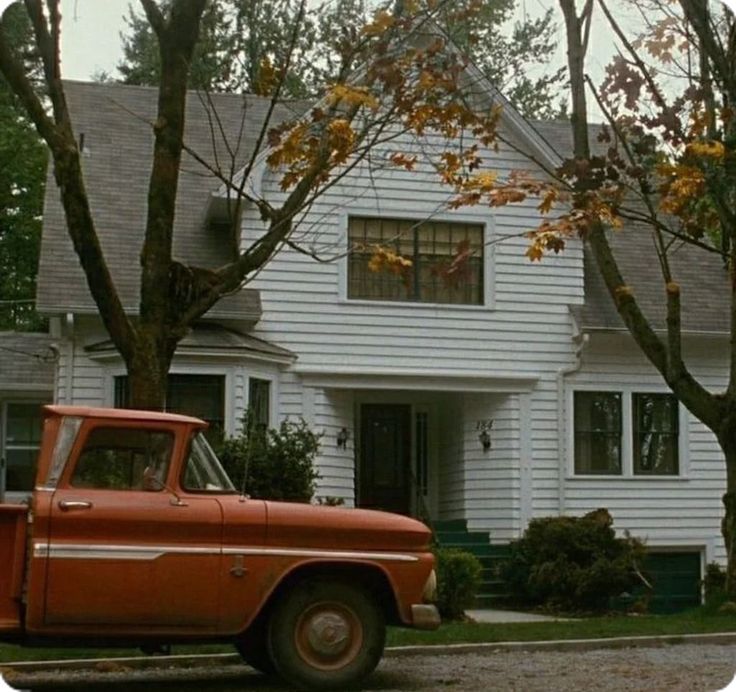 an old red truck is parked in front of a white house with trees and bushes