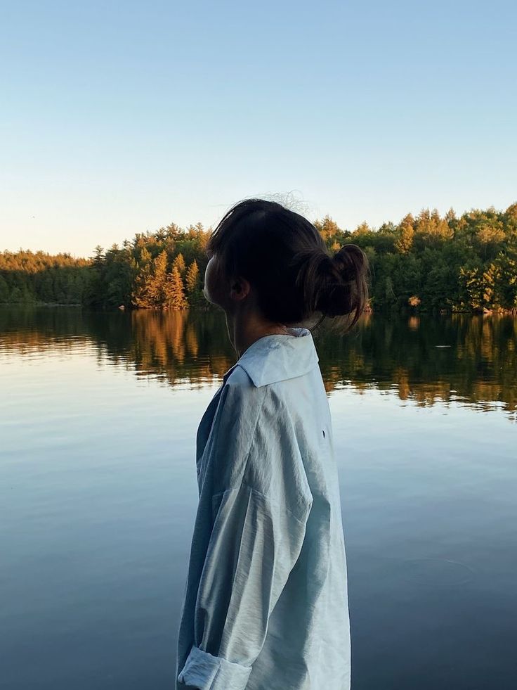 a woman standing on the edge of a body of water looking at trees in the distance