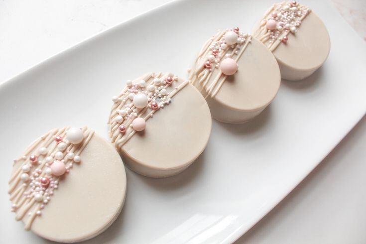 four decorated cookies on a white plate with pink beads and pearls are arranged in the shape of oval shapes