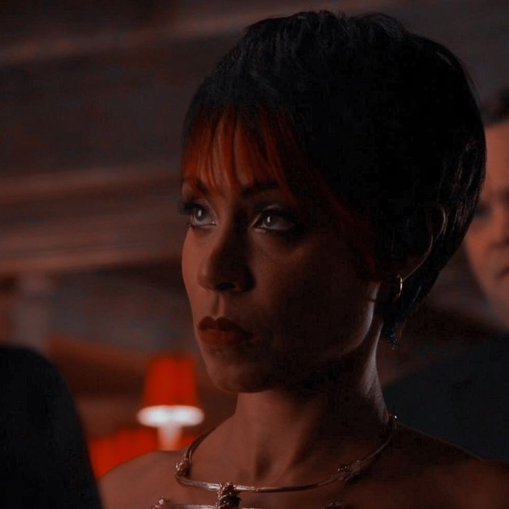 a woman with red hair wearing a necklace and earrings in a dark room next to other people