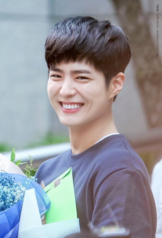 a young man holding a bouquet of flowers and smiling at the camera while standing in front of a building