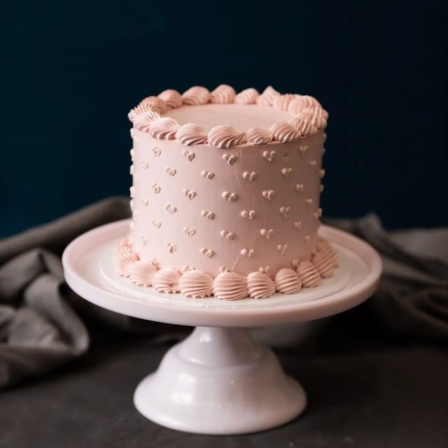 a pink frosted cake sitting on top of a white cake plate next to a gray cloth