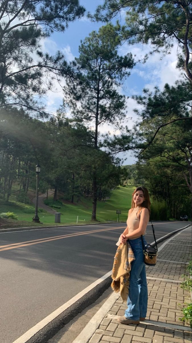 a woman standing on the side of a road with her hand in her pocket and looking off into the distance