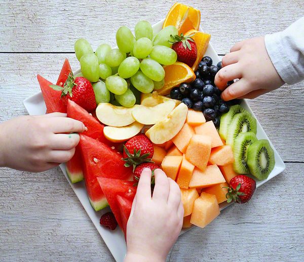 two hands reaching for slices of watermelon, oranges, and grapes on a platter