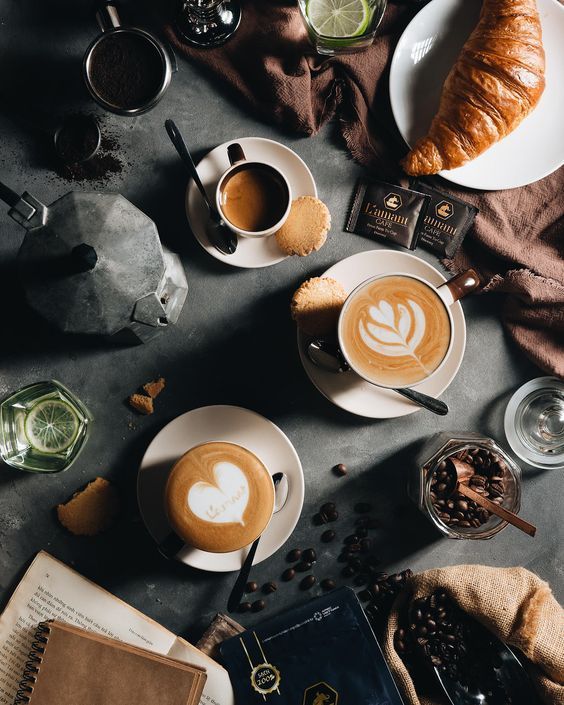 two cups of cappuccino on a table next to an open book and a croissant