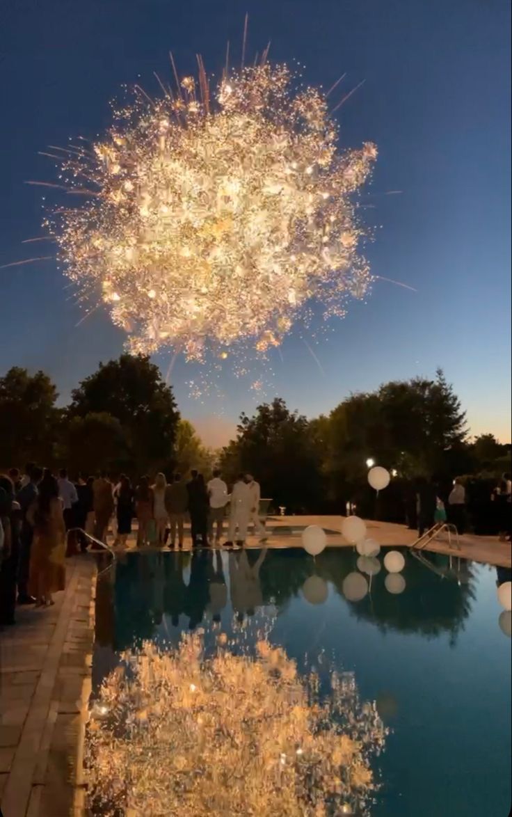 fireworks are lit up in the sky above a pool with people standing around it and balloons floating on the water