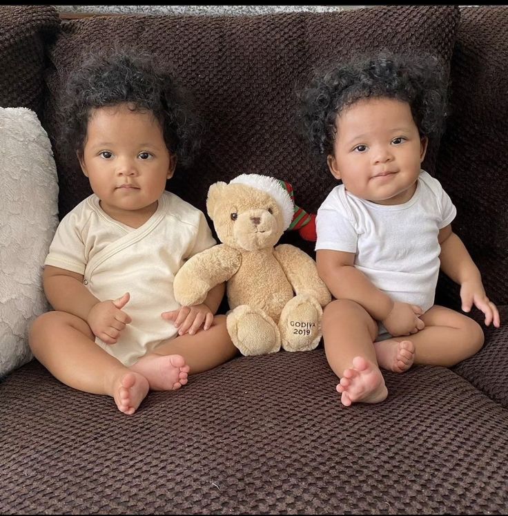 two babies sitting next to each other on a couch with a teddy bear in front of them